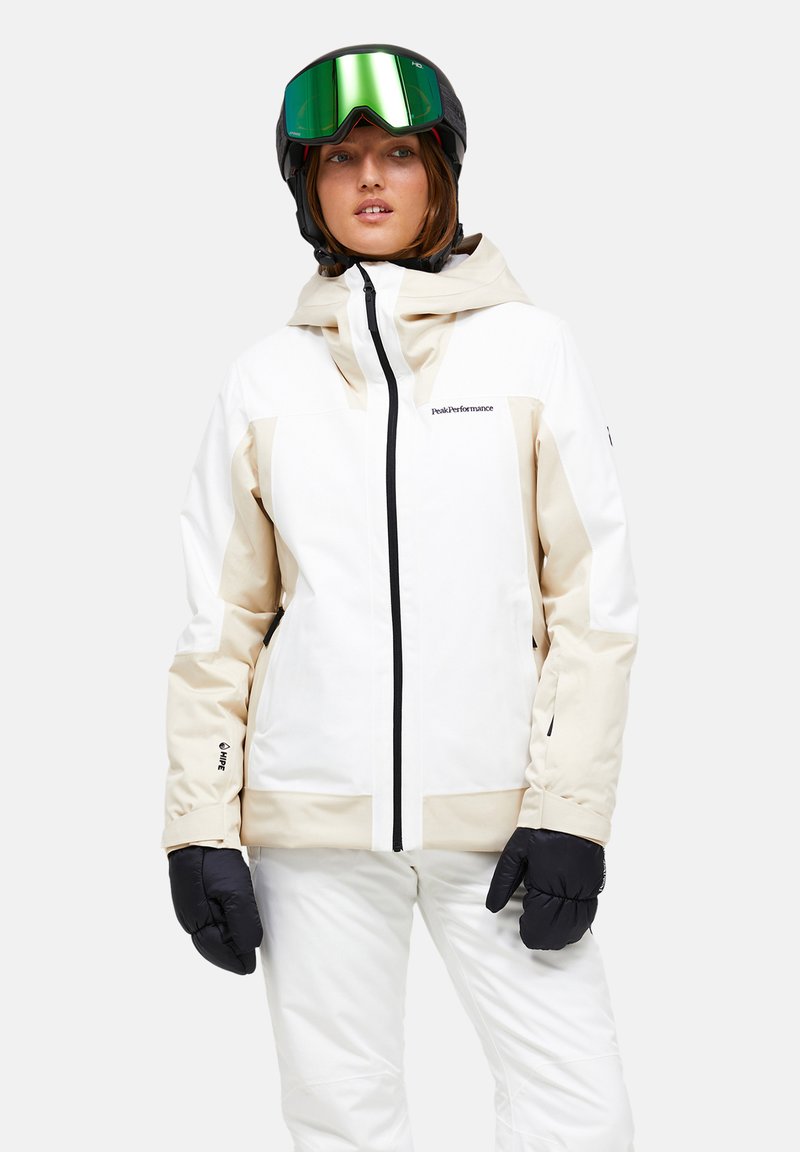 Hvid og beige skijakke med hætte, sorte lynlåse og sorte detaljer. Sammen med sorte handsker og en grøntoneret skihjelm.