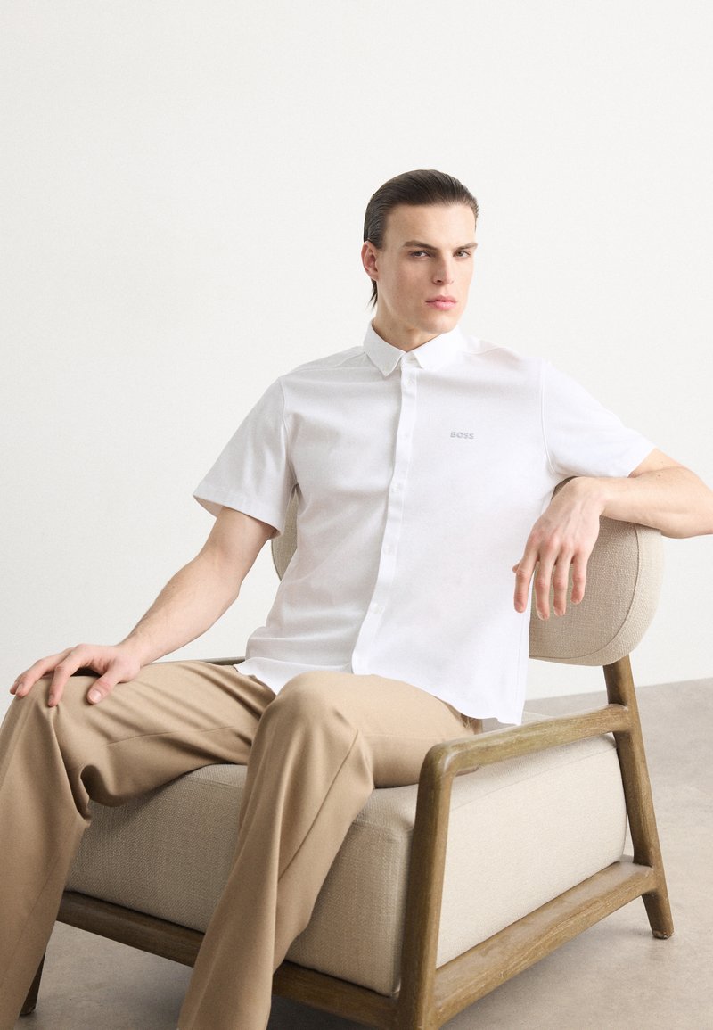 Camicia bianca a maniche corte con logo grigio, tessuto morbido, colletto classico, abbinata a pantaloni beige, seduto su una sedia di legno.