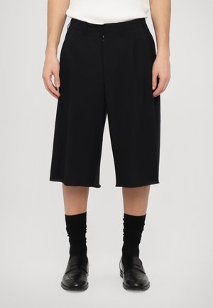 Schwarze, maßgeschneiderte Culottes mit Falten, ungesäumtem Saum und mittellangem Design. Getragen mit schwarzen Socken und Slippern, vor einem grauen Hintergrund.