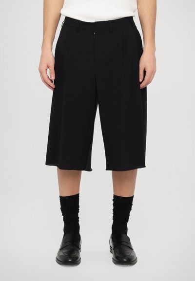 Culote negre personalizate, cu pliseuri, tiv brut și design de lungime medie. Purtați cu șosete negre și mocasini, pe un fundal gri.