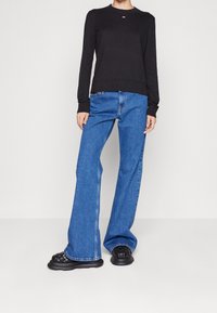 Black long-sleeve sweater with round neck, paired with blue flared jeans. Black woven slip-on shoes, casual fit, smooth fabric texture.