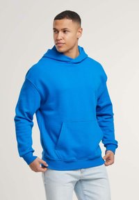 Felpa blu brillante, tessuto morbido, grande tasca a marsupio, vestibilità oversize, cappuccio regolabile con coulisse, polsini e fondo a costine.