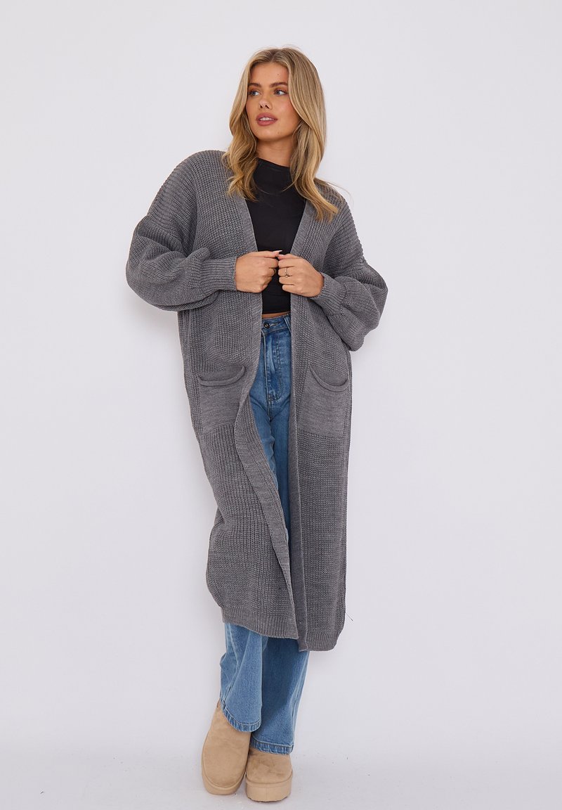 Cardigan long, ouvert sur le devant, en tricot gris texturé, avec des poches latérales, des manches larges et une coupe décontractée, associé à un jean bleu et des chaussures beiges.