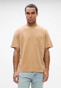 Ανοιχτό καφ�έ βαμβακερό μπλουζάκι με στρογγυλή λαιμόκοψη, κοντά μανίκια και τσέπη στο στήθος. Το ύφασμα φαίνεται μαλακό και λείο με ελάχιστες λεπτομέρειες.