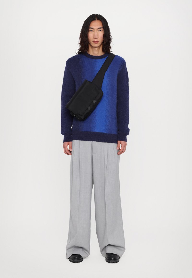 Maglione di lana a maglia con sfumature di blu, abbinato a pantaloni grigi plissettati e una borsa a tracolla nera con bottoni.
