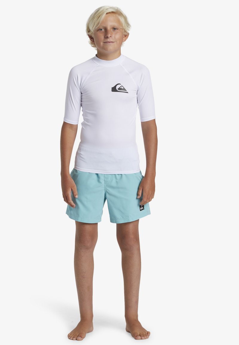 Weißes Rash Guard mit kurzen Ärmeln, versehen mit einem schwarzen Logo, kombiniert mit hellblauen Schwimmshorts. Das Model steht barfuß.