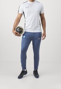 Bílá tričko s krátkým rukávem Nike s černými akcenty, modré kalhoty Nike s logem Inter Milán a černé fotbalové kopačky, které drží fotbal.