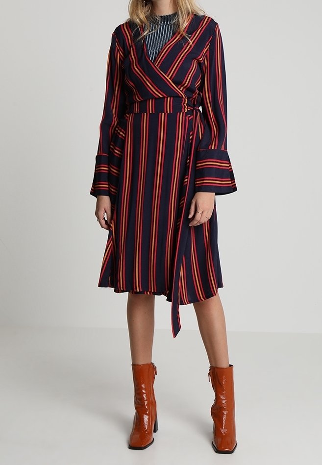 Robe portefeuille en bleu marine avec des rayures verticales rouges et oranges, dotée de longues manches évasées et d'une taille à nouer. Associée à des bottines orange brillantes.
