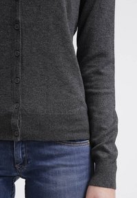 Närbild av en person som bär en mörkgrå knappad kofta och blå denimjeans, med fokus på överkroppen och högra armen.