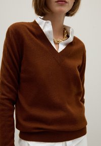 Chandail en cachemire marron avec un col en V, porté sur une chemise blanche à boutons, agrémenté d'un collier en chaîne dorée brillante.