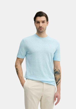 Uomo con capelli scuri che indossa una camicia azzurra a maniche corte e pantaloni beige, che mostra tatuaggi su entrambe le braccia, in piedi con una mano in tasca.