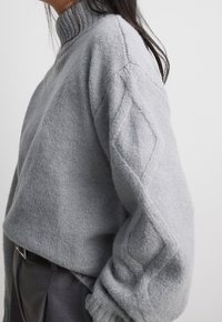 Pull tricoté gris clair avec un col montant, des manches texturées présentant un motif en losange, et des poignets côtelés. Tissu doux et coupe décontractée.