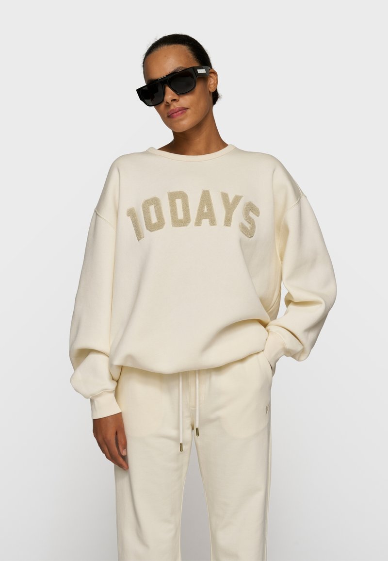 Femme portant un sweat-shirt écru oversize avec l'inscription "100 DAYS" et un pantalon de survêtement assorti, ainsi que des lunettes de soleil noires.
