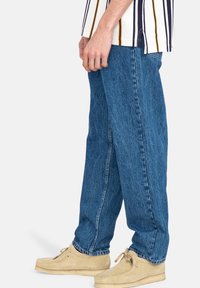 Jean en denim bleu avec une coupe ample, jambes droites et délavage clair. Assorti à des chaussures en daim beige pour un look décontracté.