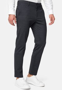 Hombre con pantalones de traje ajustados de color gris oscuro, camisa blanca y zapatillas blancas, de pie contra un fondo blanco liso.