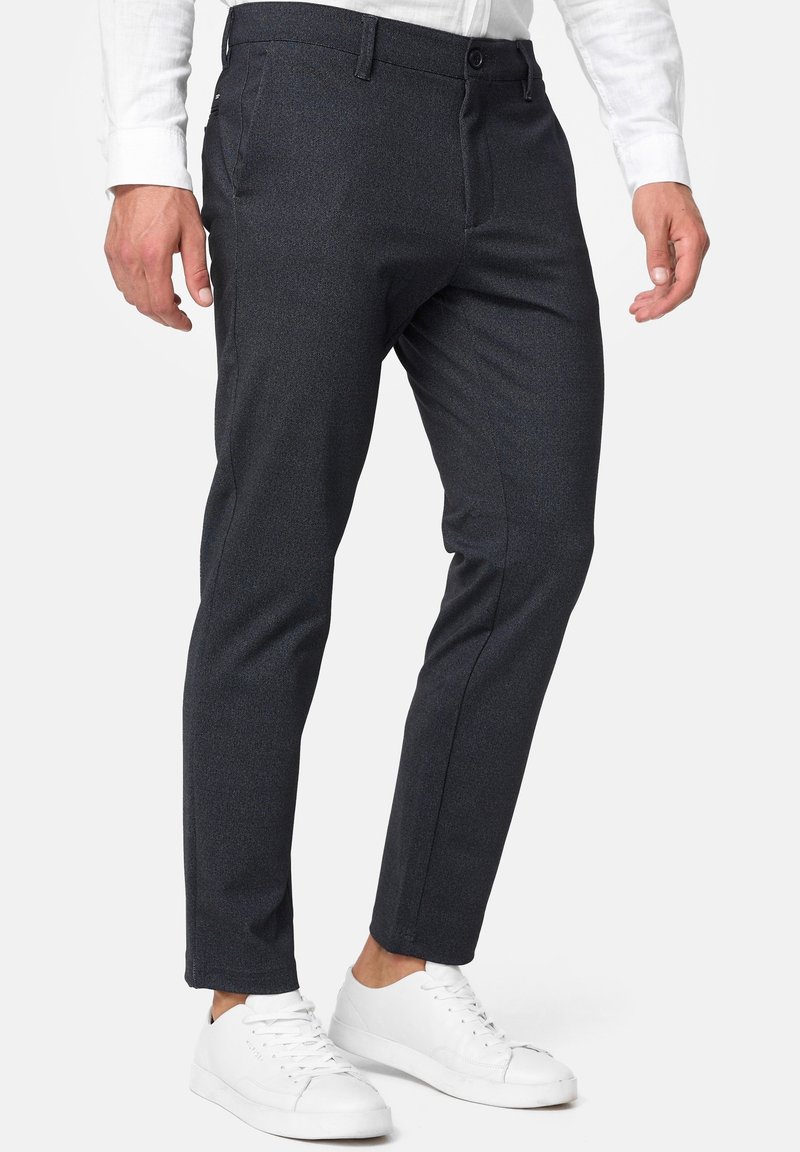 Hombre con pantalones de traje ajustados de color gris oscuro, camisa blanca y zapatillas blancas, de pie contra un fondo blanco liso.