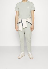 Ljusgrön t-shirt med en liten logotyp, kombinerad med matchande joggers och en krämfärgad tröja knuten i midjan. Vita sneakers med beige detaljer.