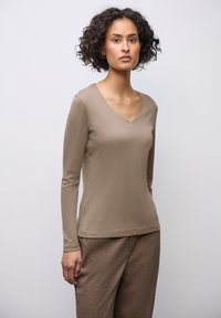 Žena s kudrnatými vlasy, oblečená v béžovém dlouhém rukávu s výstřihem do V a hnědých strukturovaných kalhotách, stojící na pozadí v jednobarevném světlém tónu.