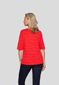 Rotes, strukturiertes T-Shirt mit Dreiviertelärmeln, das ein wellenförmiges Muster, einen runden Ausschnitt und eine lässige Passform aufweist und mit dunkelblauen Jeans kombiniert ist.
