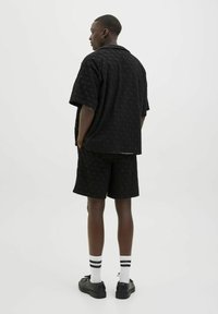 Black textured short-sleeve shirt and shorts set with a subtle pattern, paired with black shoes and striped socks. Model faces away.