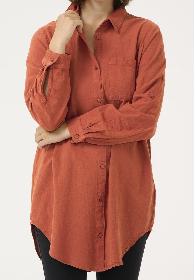 Femme portant une chemise à manches longues couleur rouille avec poche poitrine, debout les bras croisés et une main touchant le col.