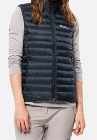 Námořnická modrá péřová vesta se zipovým uzávěrem, vertikálně štepována, s logem na levé hrudi. Vesta je spárována s světle šedým topem s dlouhým rukávem.