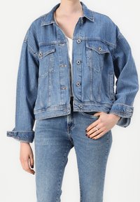 Chaqueta de mezclilla azul claro con un ajuste relajado, que cuenta con dos bolsillos en el pecho, cierre de botones y puños enrollados. Llevada sobre una camiseta blanca.