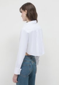 Donna con capelli castani che indossa orecchini a cerchio dorati, una maglietta bianca cropped e jeans blue a vita alta, rivolta a sinistra su uno sfondo neutro.