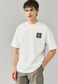 Hvid bomulds T-shirt med et firkantet grafisk design med en blomsterindretning og tekst. Korte ærmer og rund halsudskæring.