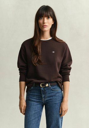 Femme aux longs cheveux bruns portant un sweat-shirt marron foncé et un jean bleu sur un fond clair uni.