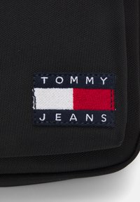 Fekete szövet táska, téglalap alakú folttal. A folton a "TOMMY" felirat található a "JEANS" fölött, piros, fehér és kék vízszintes csíkozású mintázattal.