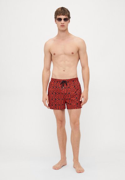 Jeune homme portant un maillot de bain à motifs rouges et noirs et des lunettes de soleil foncées, debout pieds nus sur un fond blanc uni.