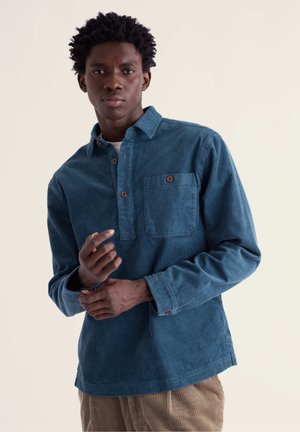 Jeune homme aux cheveux courts et bouclés portant une chemise en velours côtelé bleue et un pantalon beige, ajustant le poignet de sa chemise sur un fond uni.