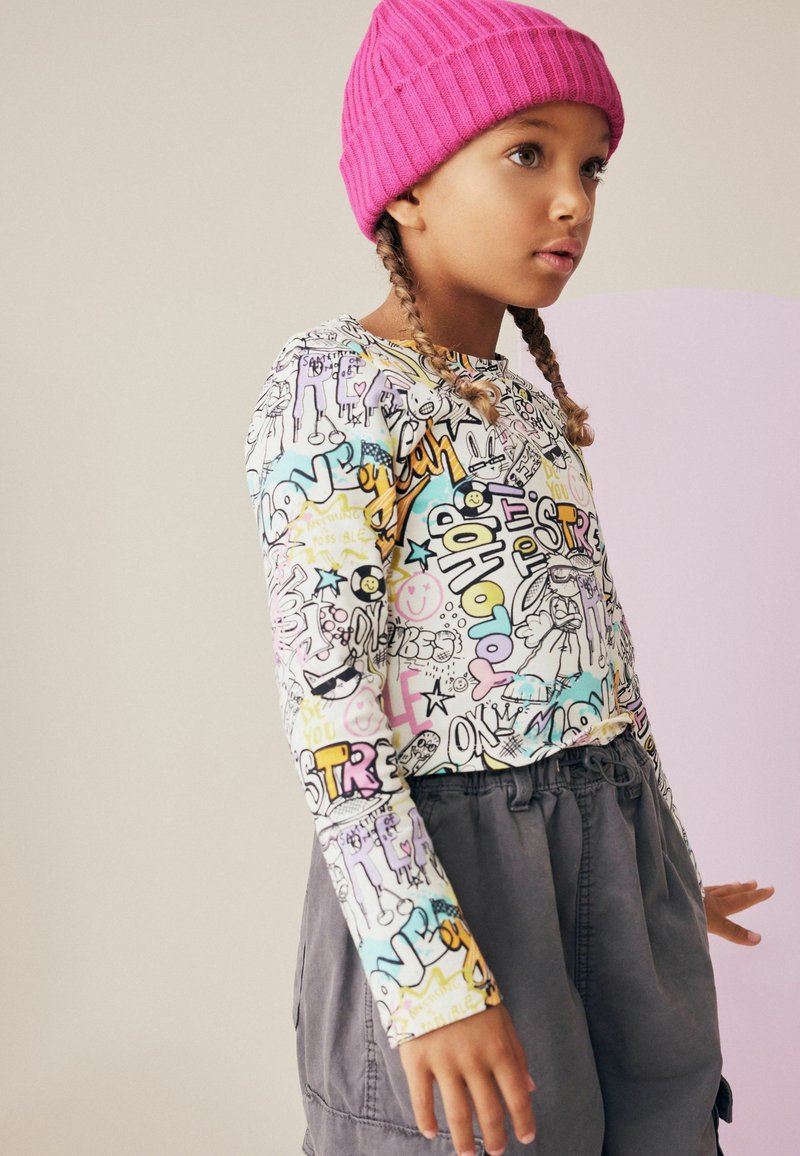 Camicia a maniche lunghe con stampa graffitata colorata in tonalità pastello, abbinata a pantaloni grigi scuri con vita elastica. Berretto in maglia di colore rosa brillante.
