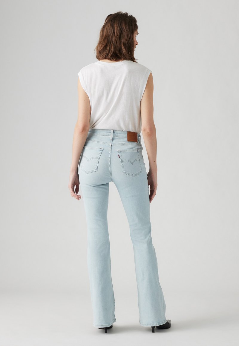 Jeans de tiro alto en azul claro, acampanados, con un parche de cuero marrón en la parte trasera, textura de denim suave y silueta ajustada.