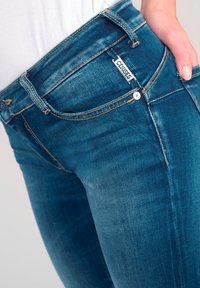 Jean en denim bleu slim avec un délavage légèrement usé, présentant des détails de couture, une étiquette de marque et une fermeture classique à bouton et fermeture éclair.