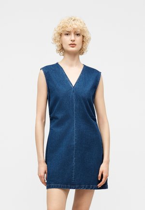 Frau mit lockigem blondem Haar, die ein ärmelloses blaues Jeanskleid mit V-Ausschnitt trägt und vor einem schlichten hellen Hintergrund steht.