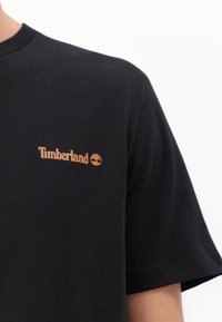 Zwart katoenen T-shirt met een klein oranje Timberland-logo op de linkerkant van de borst. Korte mouwen en een klassieke ronde halslijn.