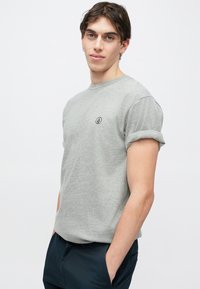 Γκρι πουκάμισο με κοντά μανίκια και στρογγυλή λαιμόκοψη, με γυρισμένα μανίκια, διαθέτει ένα μικρό μαύρο λογότυπο στο στήθος, κατασκευασμένο από μαλακό ύφασμα.