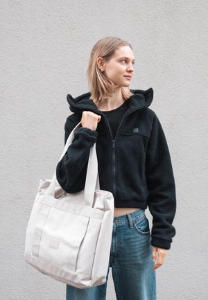 Zwarte fleece zip-up hoodie, lichtbeige schoudertas met twee hengsels, vierkante zakken en een gladde textuur, gecombineerd met blauwe spijkerbroeken.