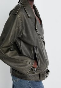 Ciemnozielona kurtka motocyklowa z imitacji skóry z asymetrycznym zamkiem, kieszeniami po bokach i szerokimi klapami. Charakteryzuje się widocznym przeszyciem i metalowymi ozdobami.