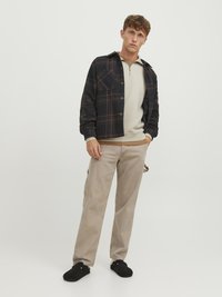Schwarz-braune kariert Jacke über einem beigen Reißverschluss-Sweatshirt, kombiniert mit hellbeigen Hosen und schwarzem Schuhwerk. Einfaches Design und Textur.