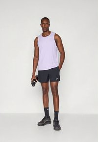 Canotta lilla chiaro, pantaloncini neri con un logo bianco, scarpe da ginnastica nere, tiene una bottiglia d'acqua, in piedi su uno sfondo semplice.