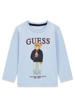 Chemise bleu clair à manches longues avec un ours portant un jean et un sweat à capuche noir, logo "GUESS" en haut et "Original Authentic Brand Est. 1981" en bas.
