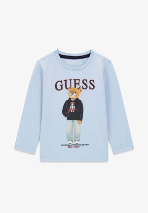 Jasnoniebieska koszula z długim rękawem z niedźwiedziem w dżinsach i czarnej bluzie z kapturem, logo "GUESS" powyżej i "Original Authentic Brand Est. 1981" poniżej.