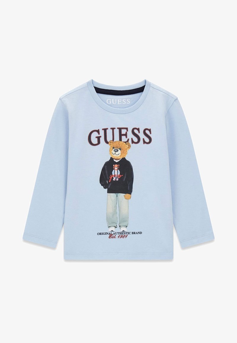 Chemise bleu clair à manches longues avec un ours portant un jean et un sweat à capuche noir, logo "GUESS" en haut et "Original Authentic Brand Est. 1981" en bas.