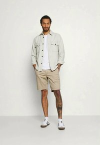 Calções bege com uma t-shirt branca por baixo de uma camisa abotoada cinza-claro. O modelo usa sapatilhas brancas com riscas pretas e tem tatuagens visíveis.