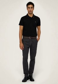 Czarna koszulka polo z kołnierzykiem, szare dopasowane spodnie i czarne botki za kostkę. Model stoi prosto, ręce w kieszeniach, neutralne tło.