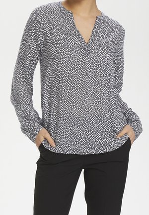 Femme portant un chemisier noir à manches longues avec un petit motif floral blanc et un pantalon noir, les mains dans les poches, sur un fond uni.
