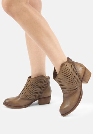 Ben iført brune læderankelstøvler med perforerede bølgemønstre og lave, stablede træsko på hvid baggrund.